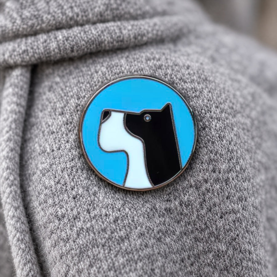 Blue circular enamel pin with a dog and cat silhouette on grey fabric
