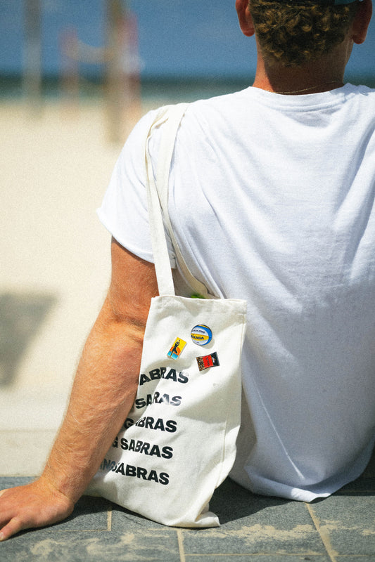 Person wearing a white t-shirt with a tote bag featuring logos and pins, sitting on a beach.