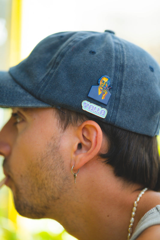 Close-up of a person wearing a blue cap with a yalla and gold pin, outdoors.