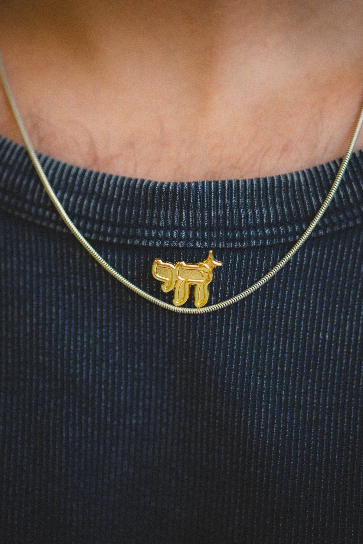 Gold necklace with a chai-shaped pin on a person wearing a dark blue shirt.