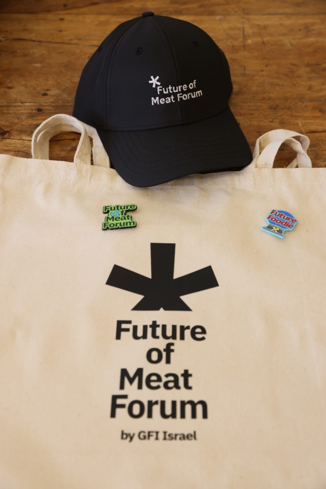Black cap and beige tote bag with 'Future of Meat Forum' branding on a wooden surface.