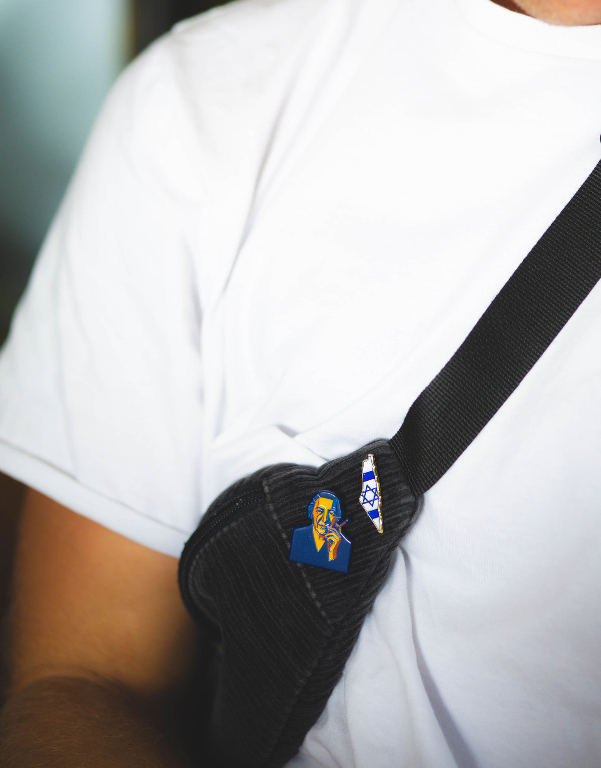 Person wearing a white shirt with a black strap over one shoulder, featuring two pins.