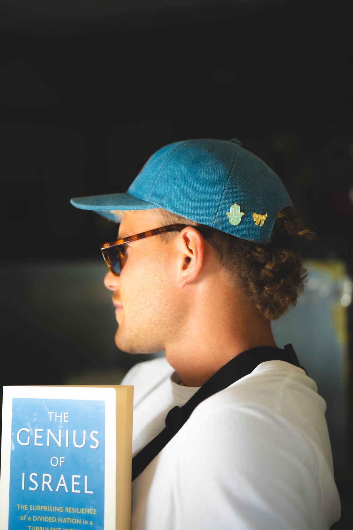 Man wearing a blue cap and sunglasses, holding a book titled 'The Genius of Israel'.