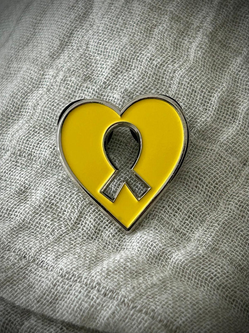 Yellow heart-shaped pin with a silver ribbon symbol on a textured gray background